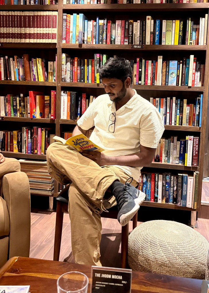 Jayesh Bhaware Reading Book in a library. Sitting on chair, legs crossed.